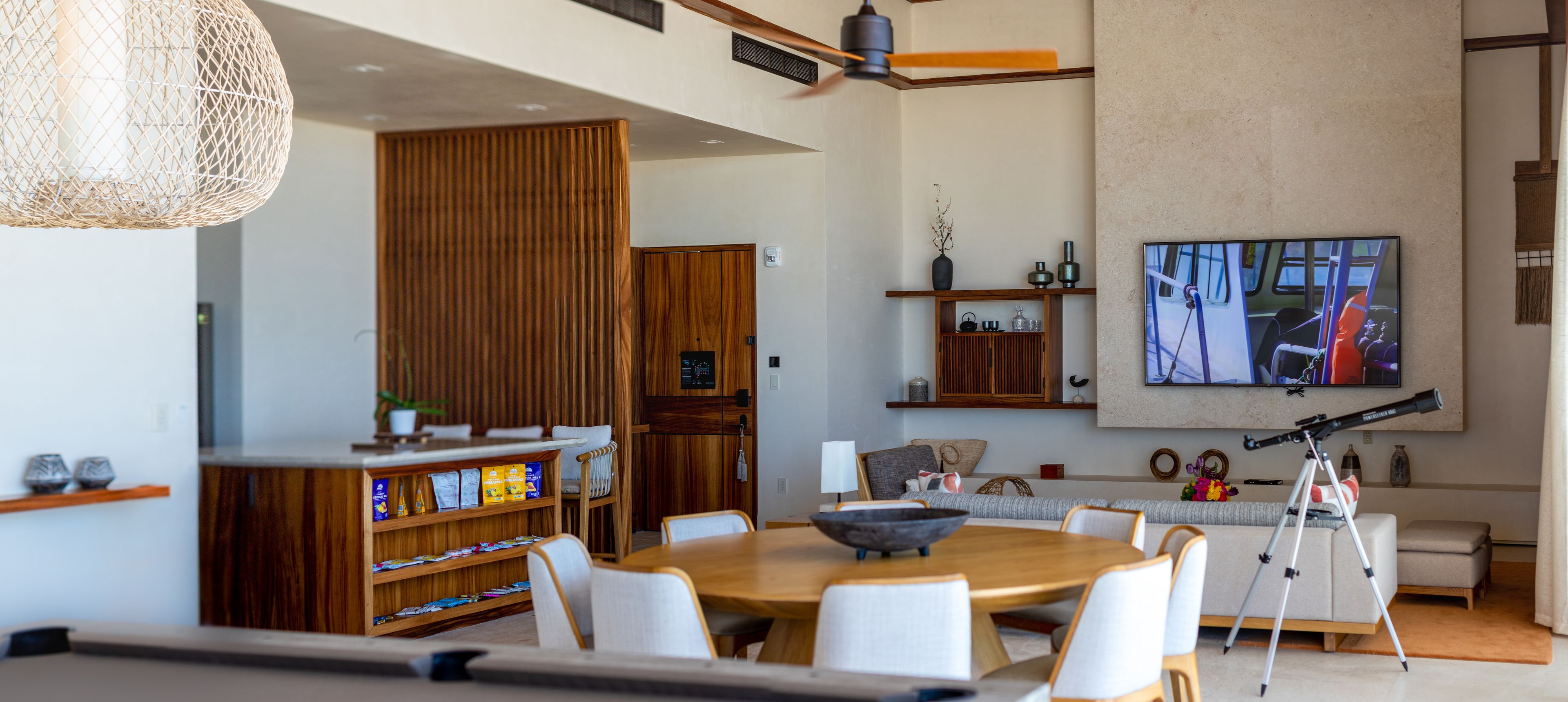 A modern living room with a dining area, wooden furniture, a bookshelf, a telescope, and a flat-screen TV mounted on the wall.
