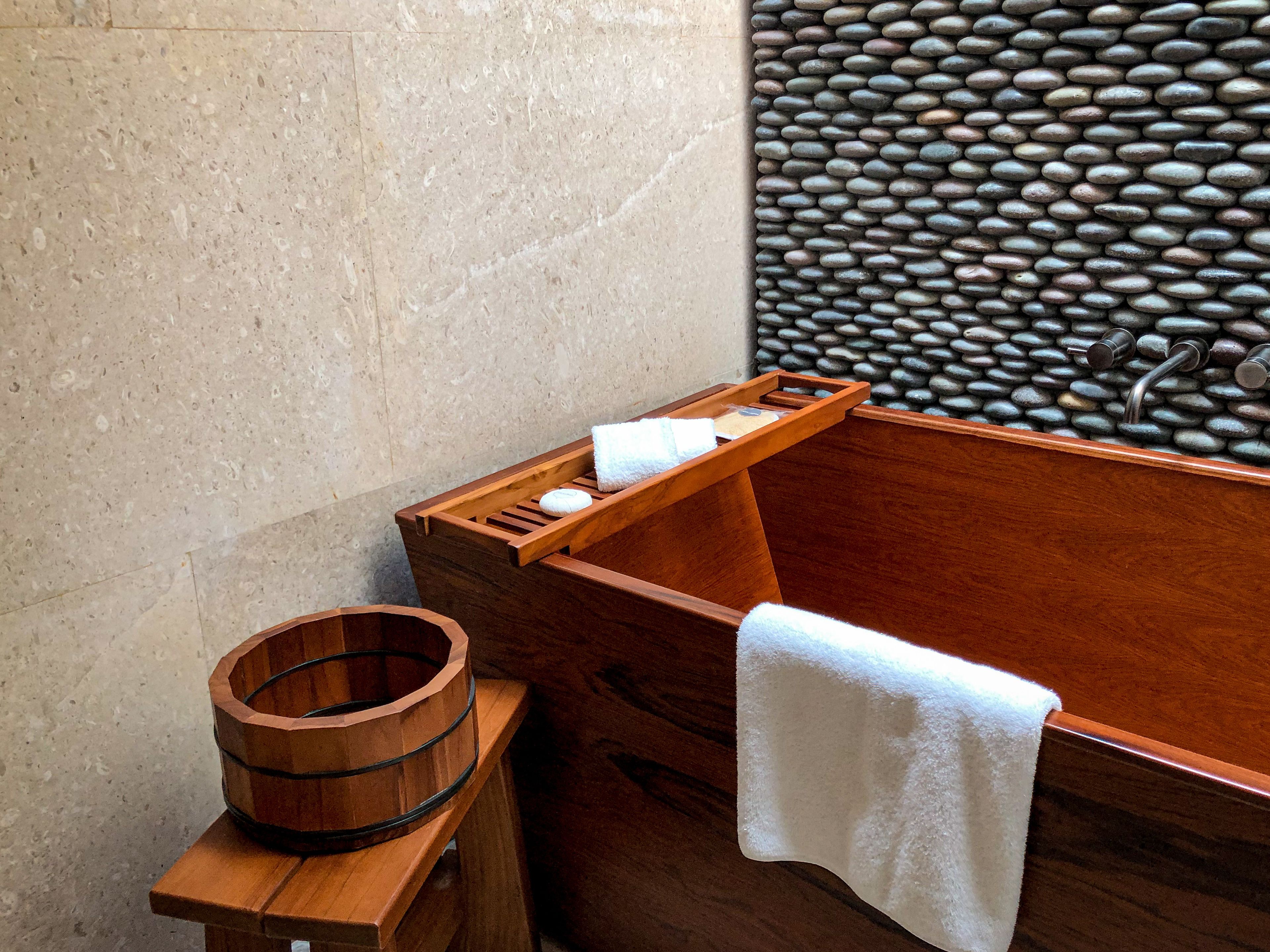 Une baignoire en bois, avec un mur en pierre en arrière-plan, un seau en bois et une serviette posée sur un tabouret en bois.