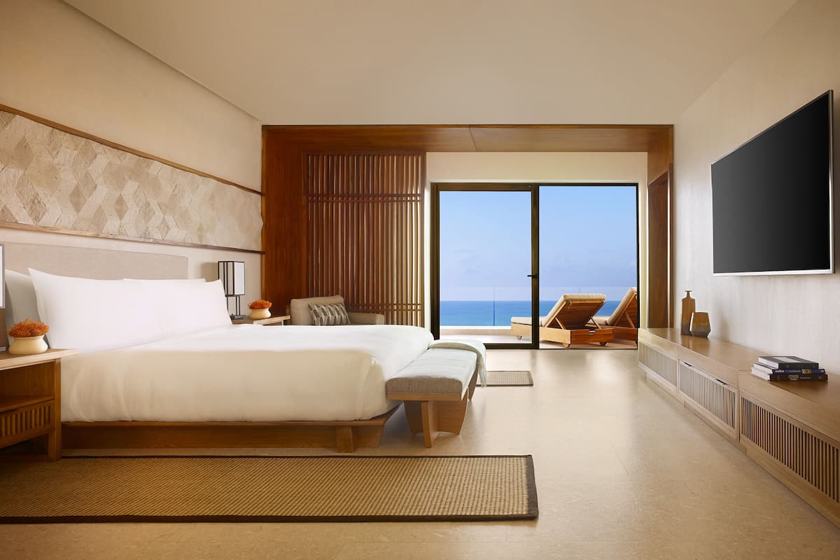 A modern hotel room with a large bed, a TV, a view of the ocean, and a balcony with a lounge chair.