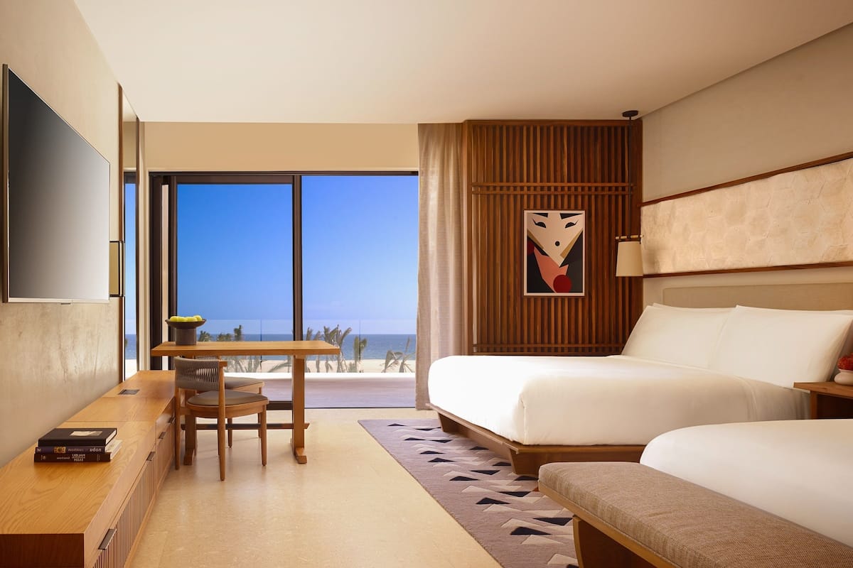 A hotel room with a bed, a desk, and a chair. There is a large window with a view of the beach.