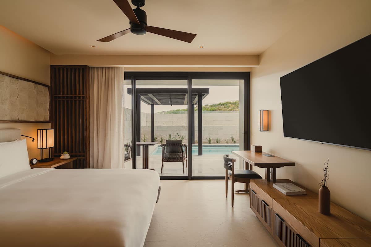 A hotel room with a large bed, a desk with a chair, a ceiling fan, and a view of a pool and greenery outside.