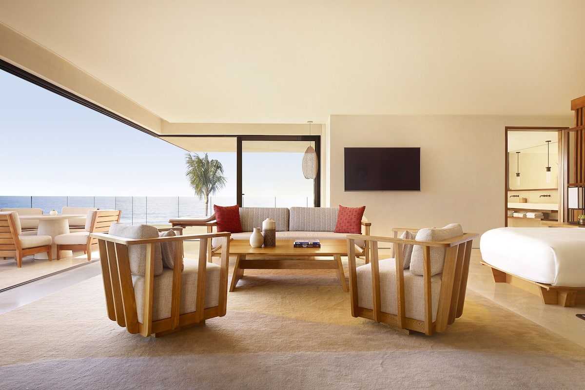 A modern living room with a large window overlooking the sea, furnished with wooden furniture and a flat-screen TV mounted on the wall.