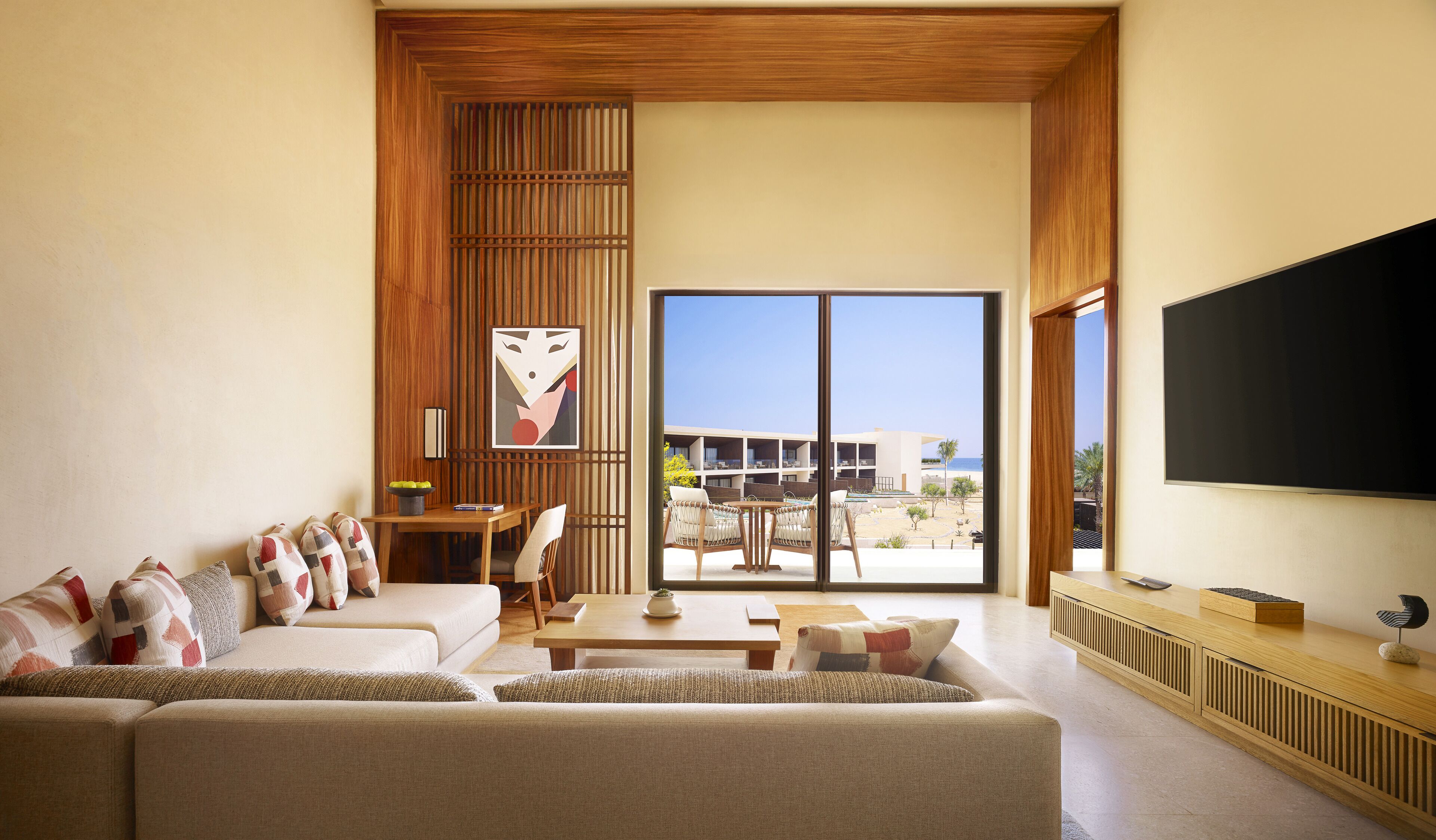 Amplia sala de estar con un sofá grande, una mesa de centro y un televisor montado en la pared. Una puerta corrediza de vidrio da a un área exterior con piscina y un edificio al fondo.
