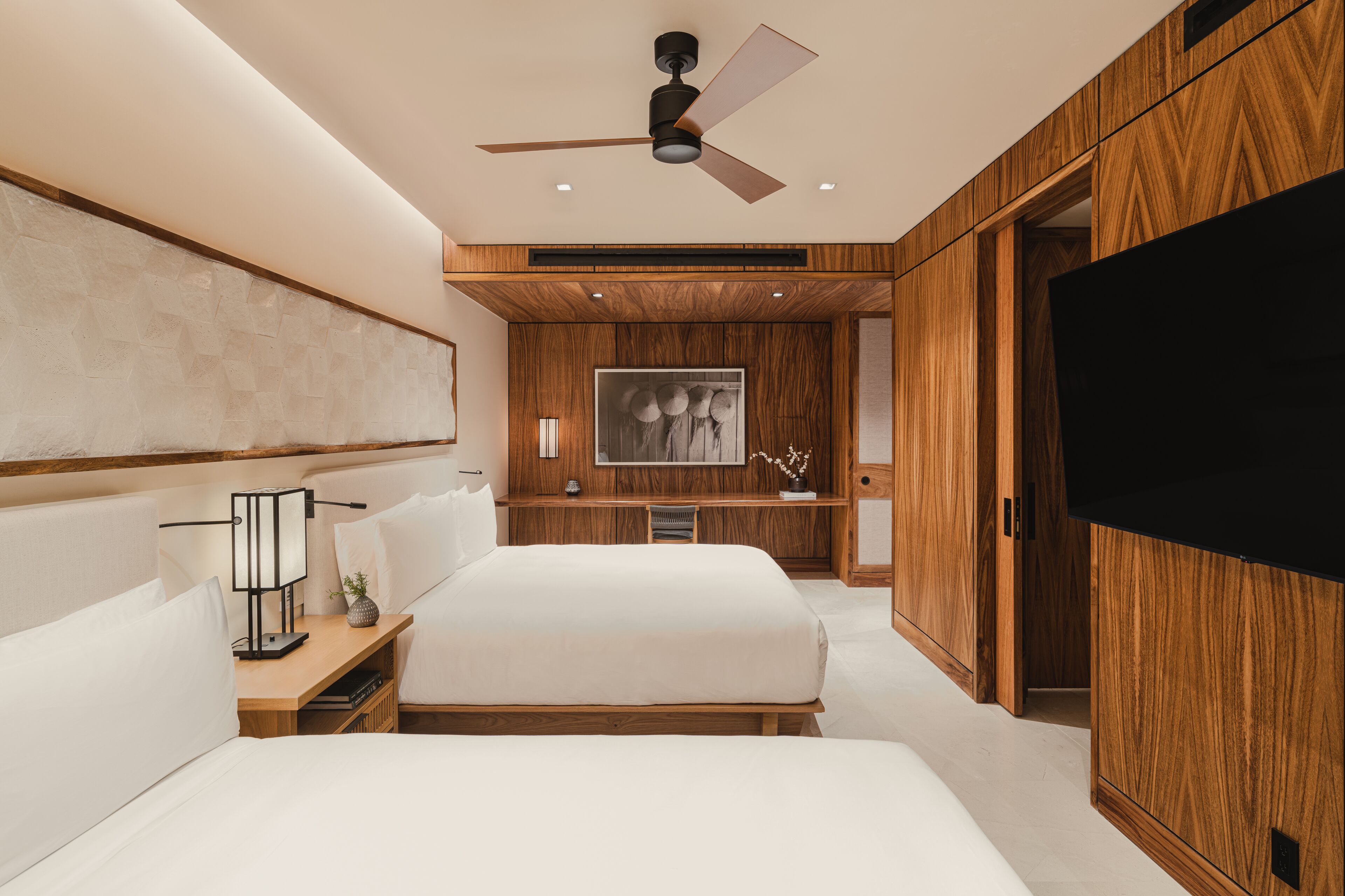 A modern bedroom with a large bed, wooden paneling, a ceiling fan, a flat-screen TV, and a desk with a lamp.