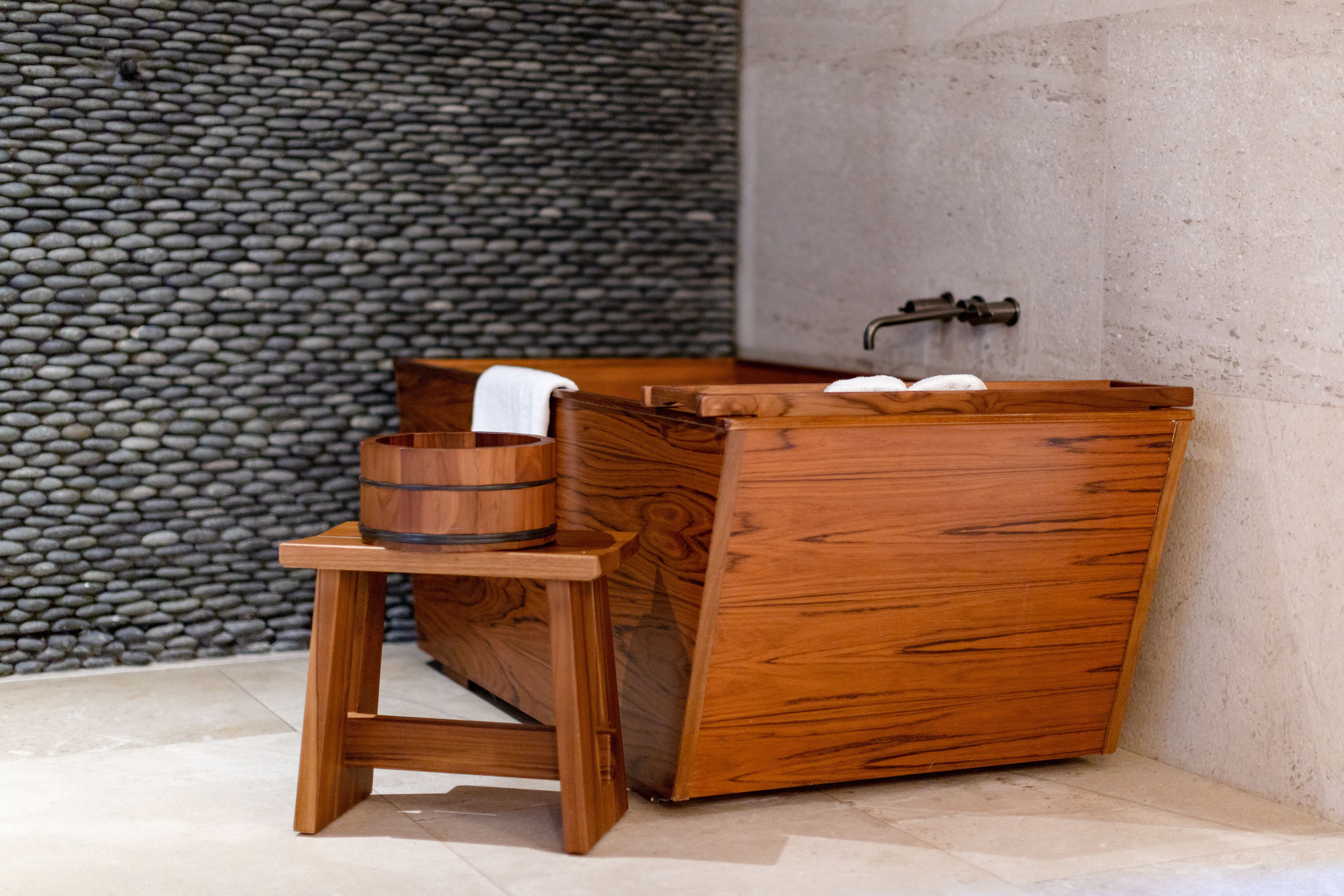 A wooden bathtub with a wooden stool beside it, set against a stone wall.