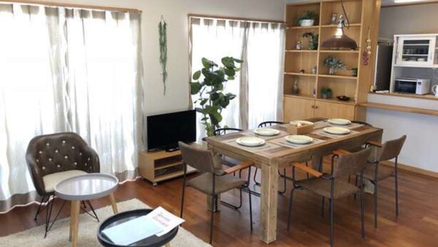 A dining area with a wooden table set for four, surrounded by chairs. There is a television on a stand to the left, and a bookshelf with various items to the right. - Villa Sans Company (Ishigaki)