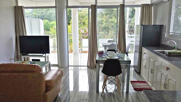 A modern living room with a sofa, a dining table with chairs, a TV, and a kitchen area with a refrigerator and sink.
