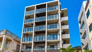 Un edificio de varios pisos con balcones, un escaparate visible con señalización y palmeras cercanas.