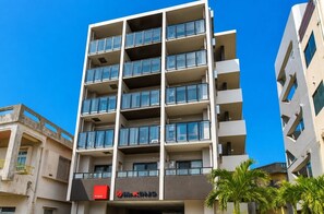 Un immeuble à plusieurs étages doté de balcons, une devanture visible avec une enseigne, et des palmiers adjacents.