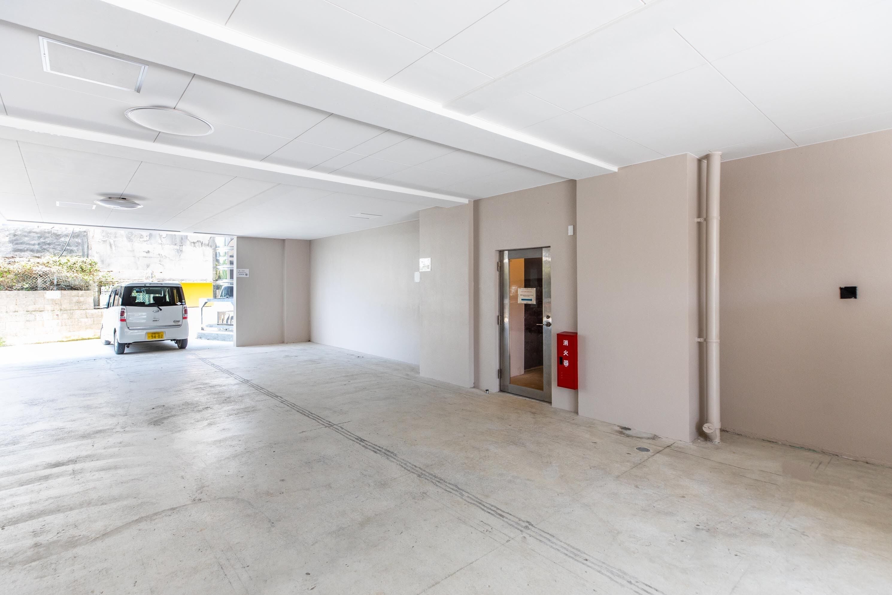 Un parking avec une voiture blanche garée au loin, un boîtier d’alarme incendie et une porte avec une pancarte.