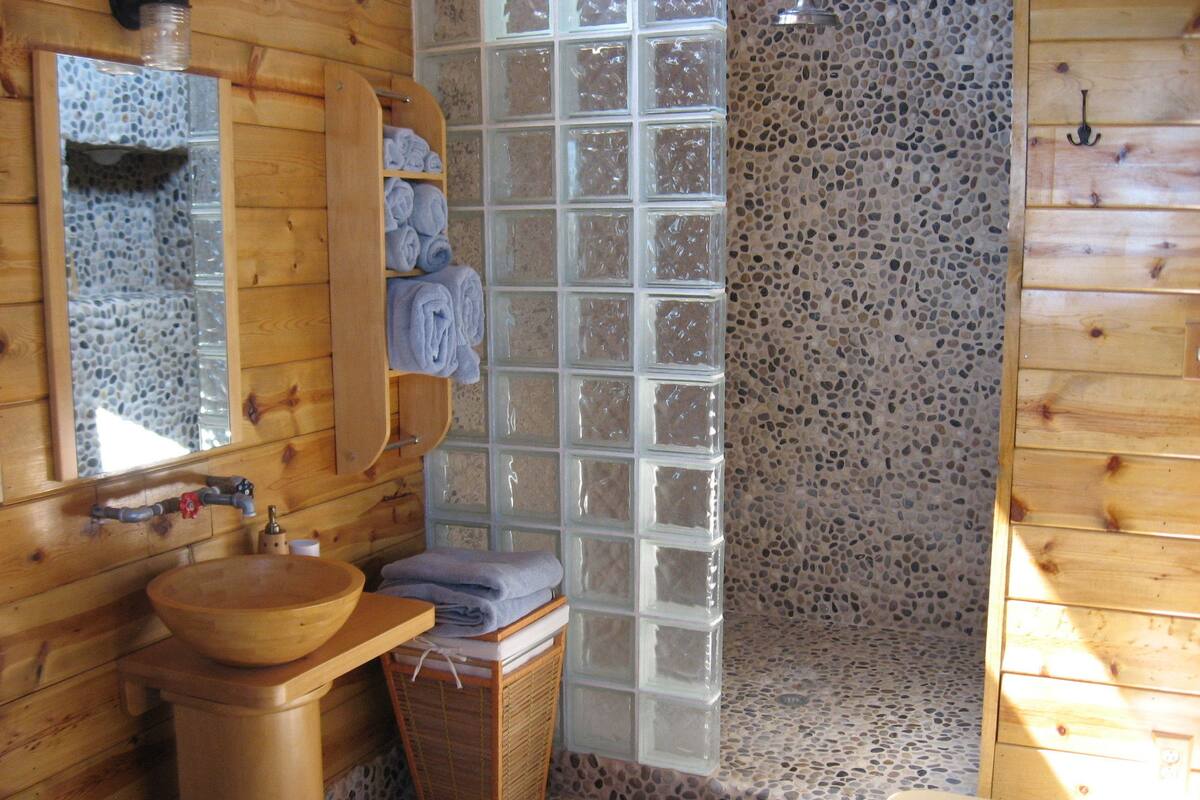 Un baño de madera con lavamanos de madera, un espejo, un perchero para toallas y una ducha con pared de guijarros.
