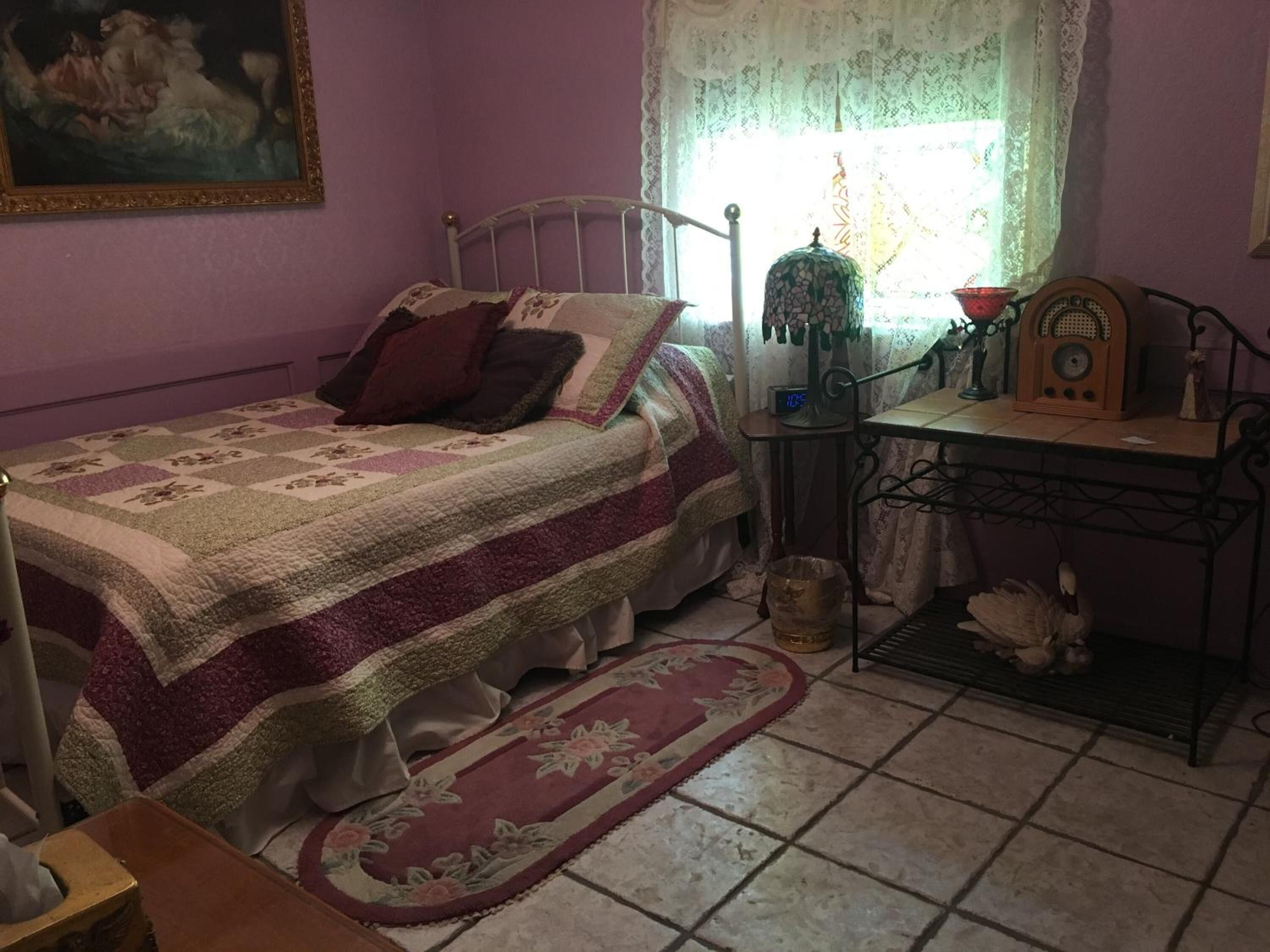 A bedroom with a bed, a nightstand, a radio, and a window with lace curtains.
