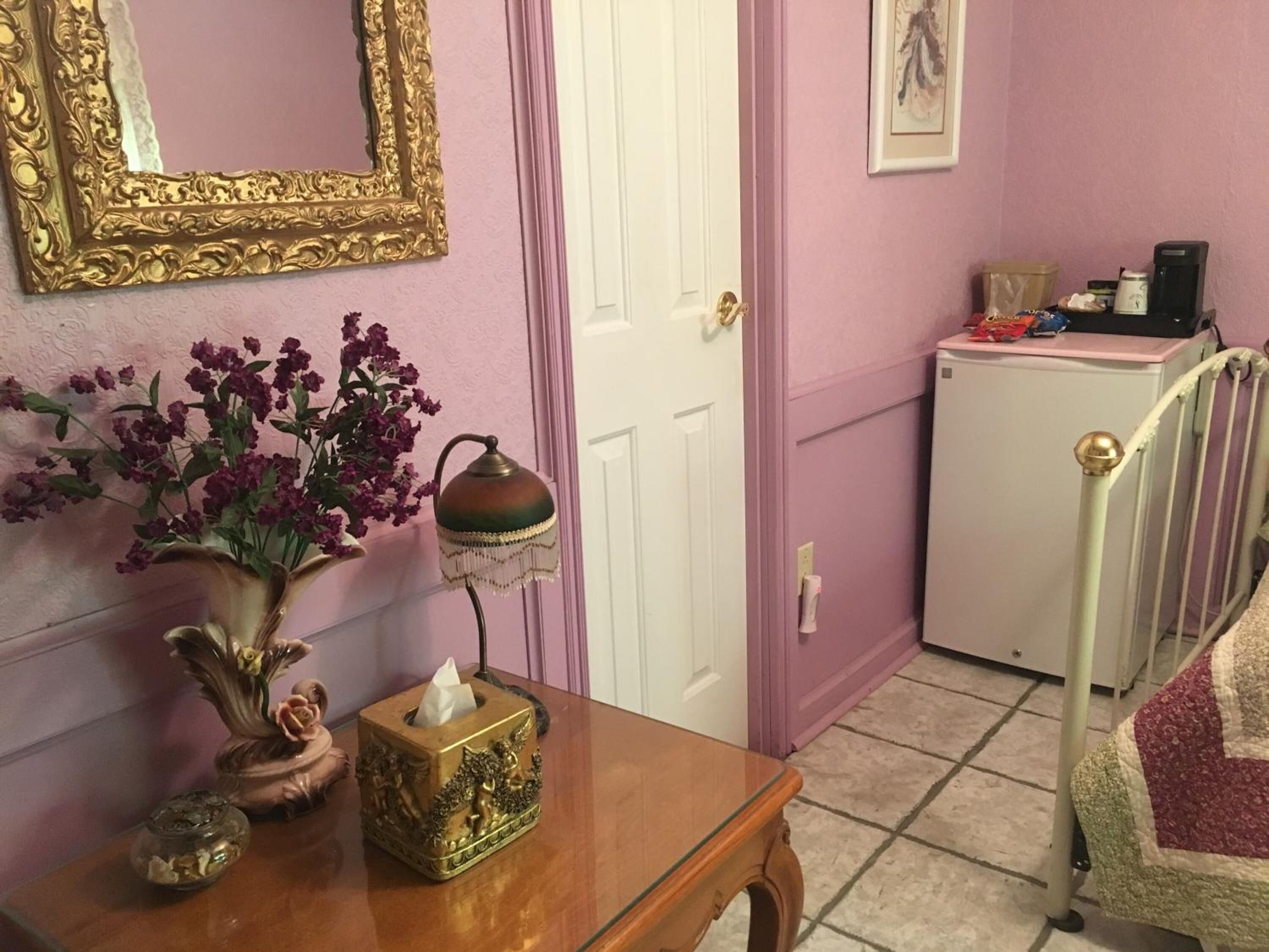 A room with a wooden table, a vase with flowers, a tissue box, a lamp, a door, a bed with a quilt, and a small refrigerator.
