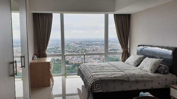 A bedroom with a large bed, a nightstand, and a view of a cityscape through a large window.