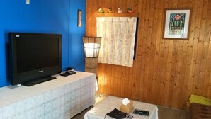 Una habitación con una televisión sobre un mueble blanco, una pequeña mesa con un control remoto, y una pared de madera con una cruz y obras de arte.