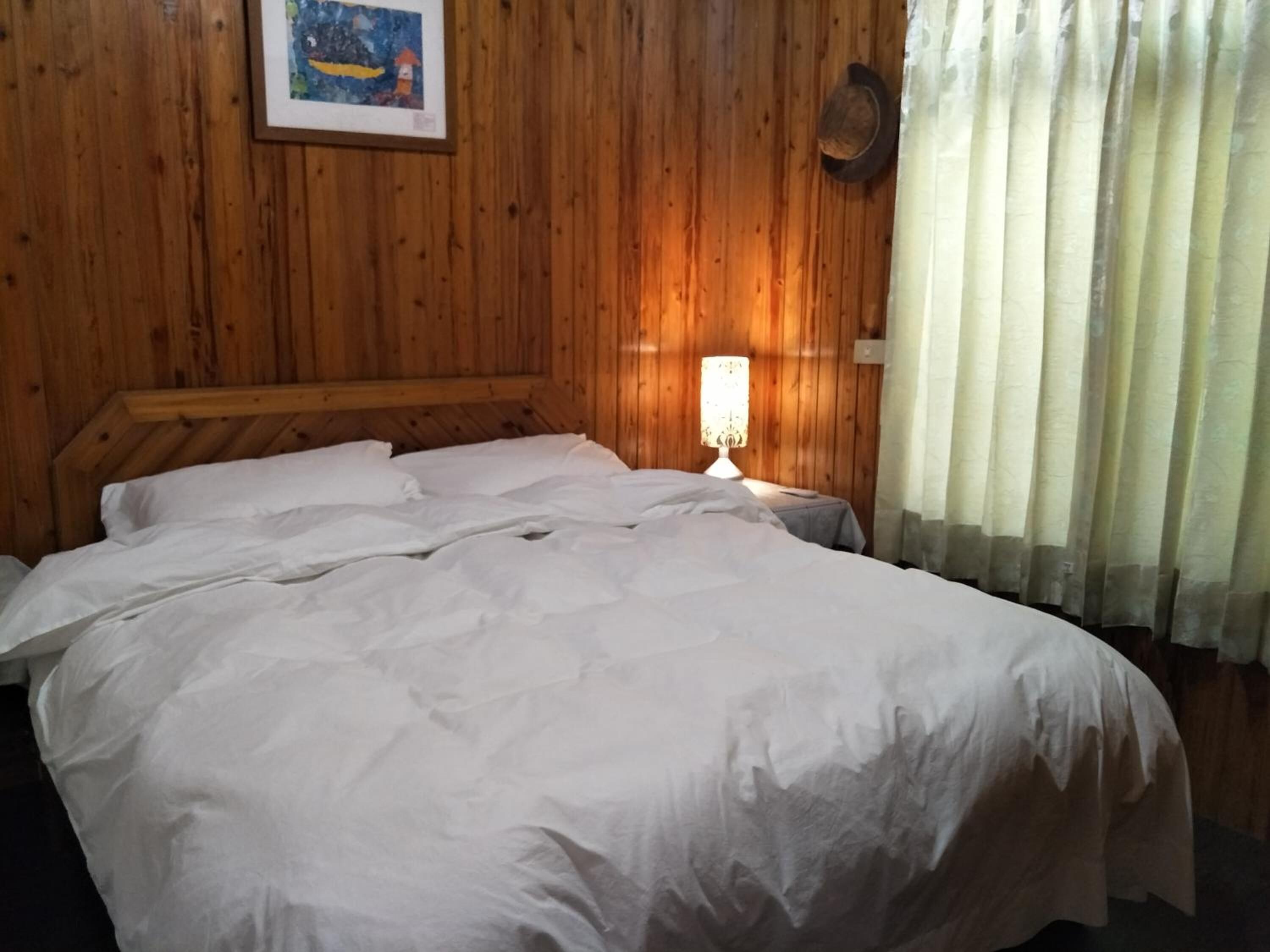 Een kamer met houten lambrisering, een bed, nachtkastje, lamp en een schilderij aan de muur.