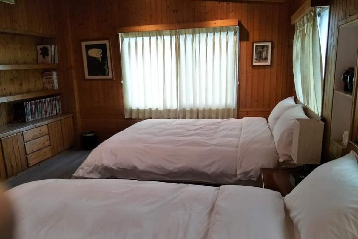 A room with two beds, wooden walls, a window with curtains, and a bookshelf with books and DVDs.