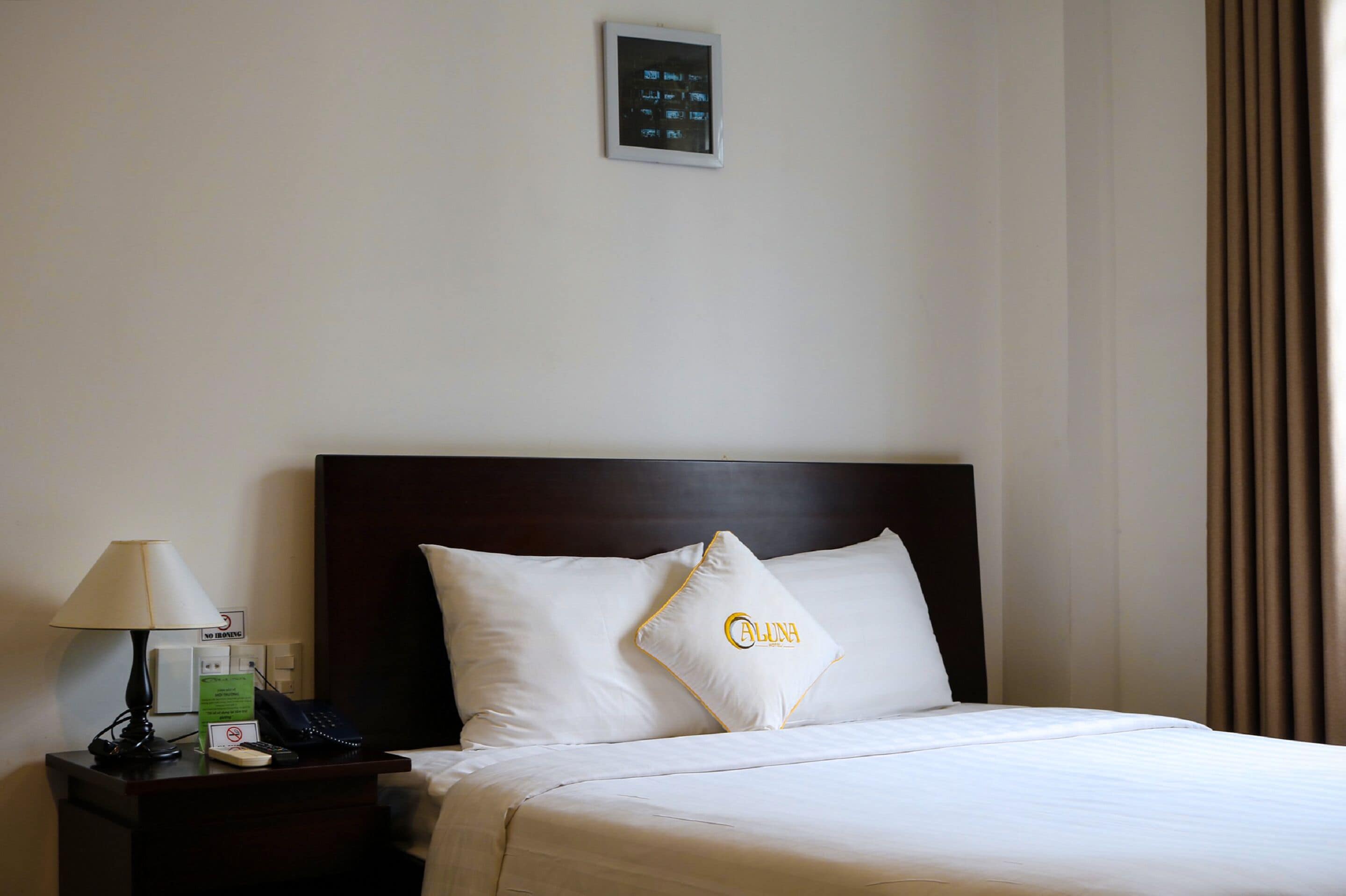 A hotel room with a neatly made bed, a bedside table with a lamp, a phone, and a sign.