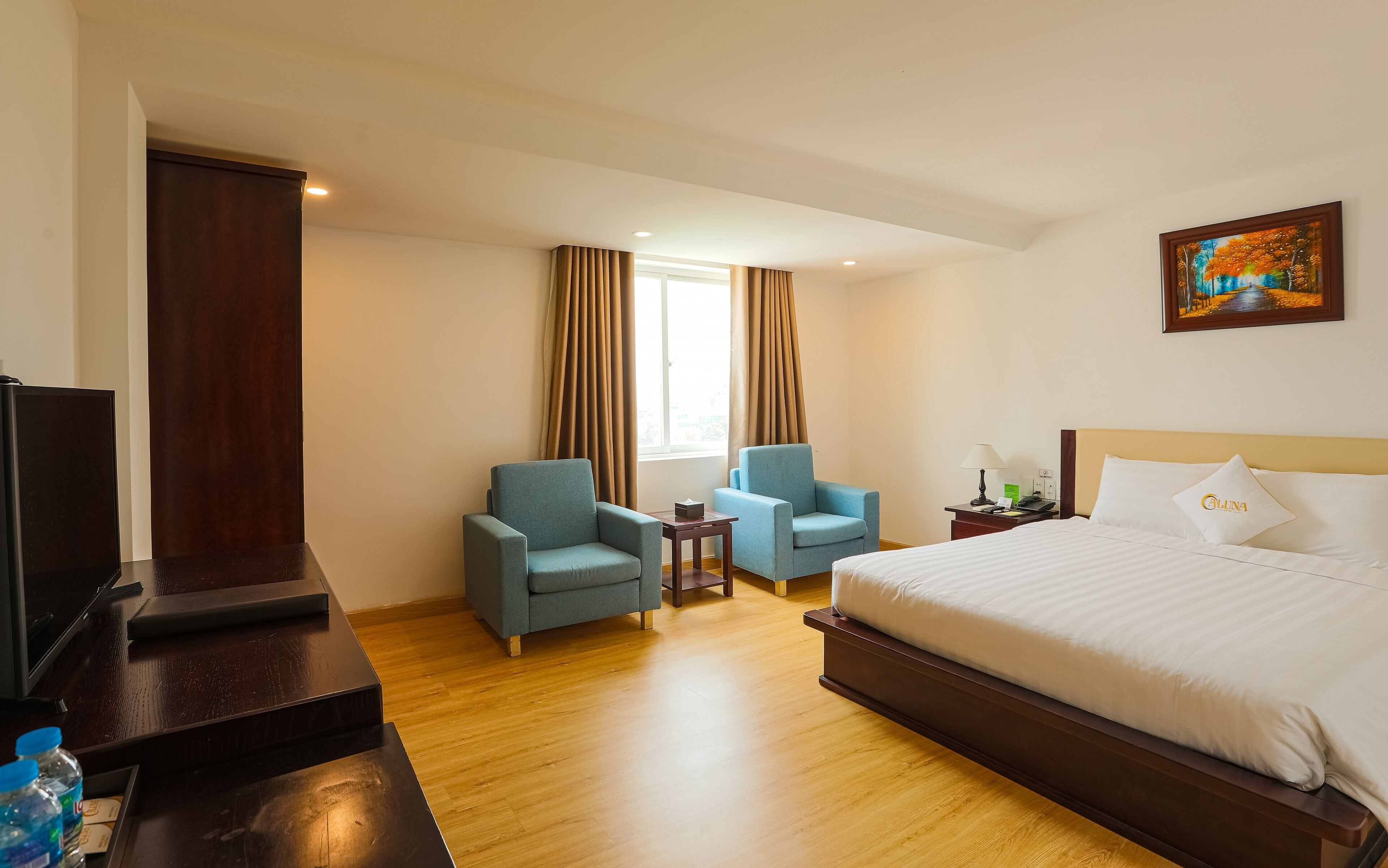 Una habitación de hotel con cama, dos sillones, un escritorio con computadora, una ventana con cortinas y un cuadro en la pared.