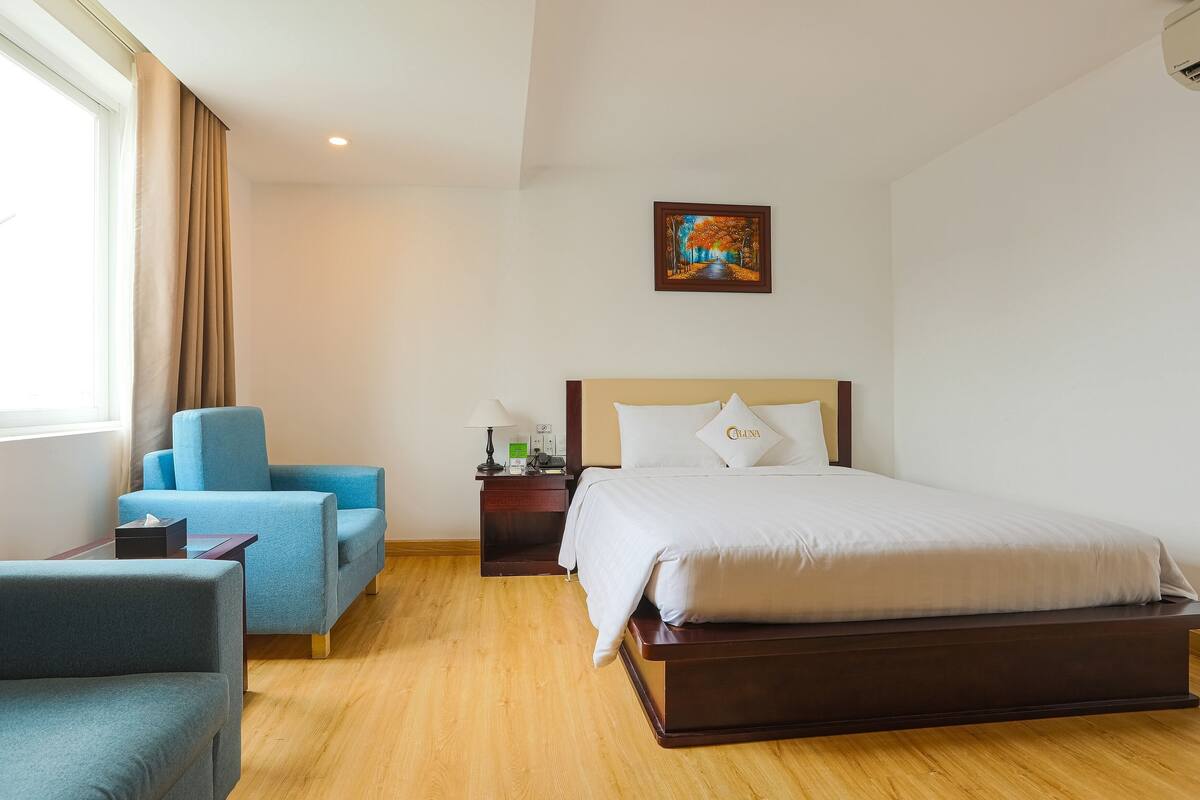 Habitación de hotel con una cama grande, un sillón azul, una mesita de noche de madera y un cuadro en la pared.