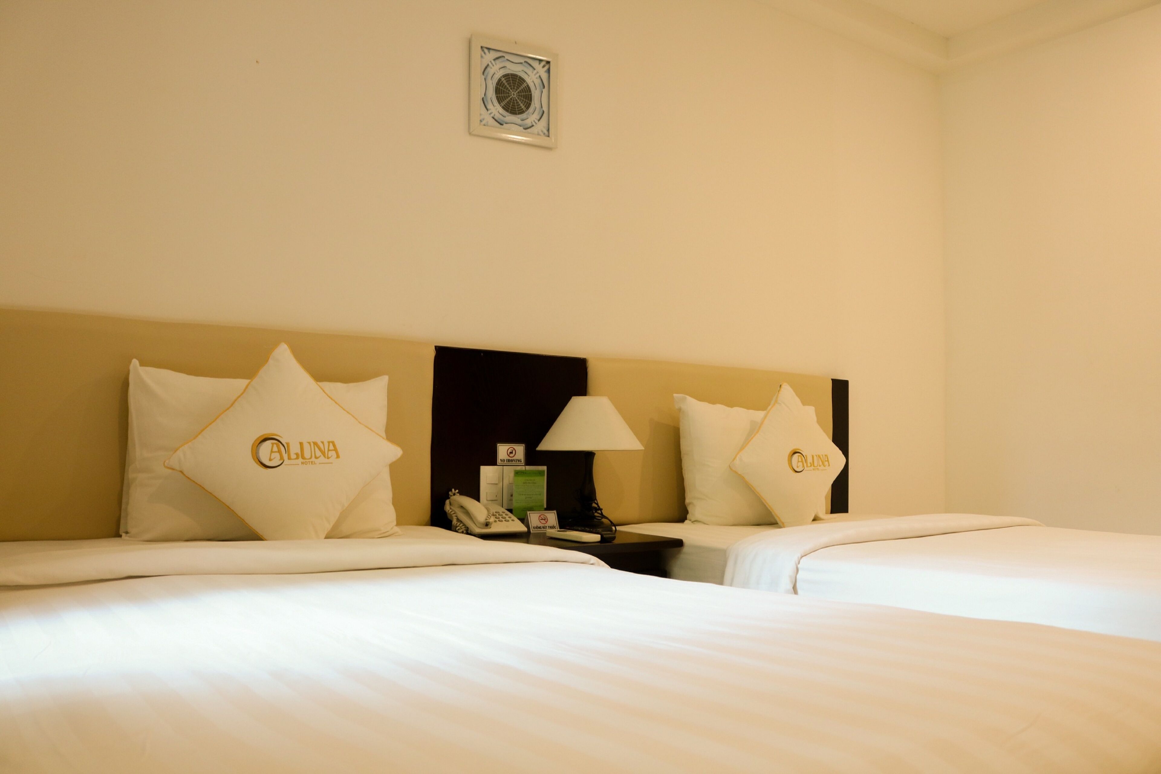 A hotel room with two beds, each with a pillow featuring the hotel's logo. There is a nightstand with a lamp, a phone, and a sign indicating no smoking.