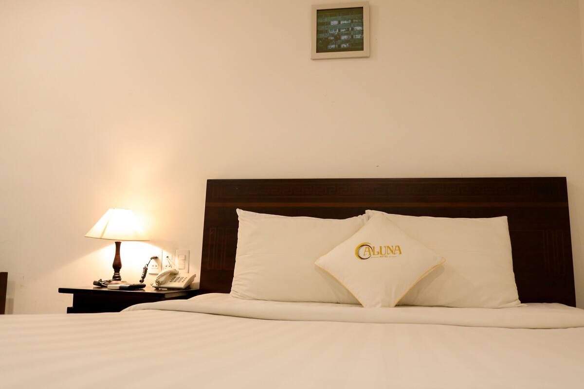 Una habitación de hotel con una cama bien hecha, una lámpara de noche, un teléfono y una pantalla en la pared.