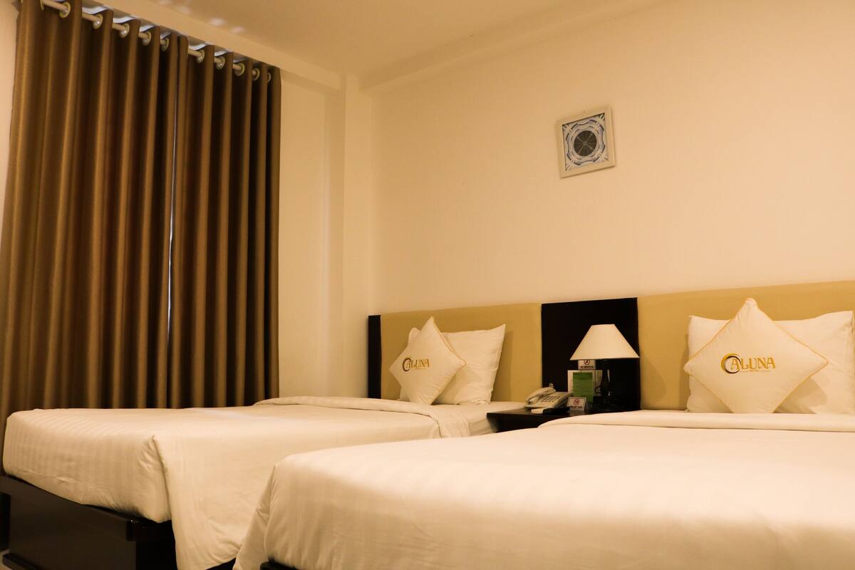 Habitación de hotel con dos camas, cada una con cubrecama y almohadas que muestran el nombre de la marca. Se observa un mesita de noche con una lámpara y un teléfono.