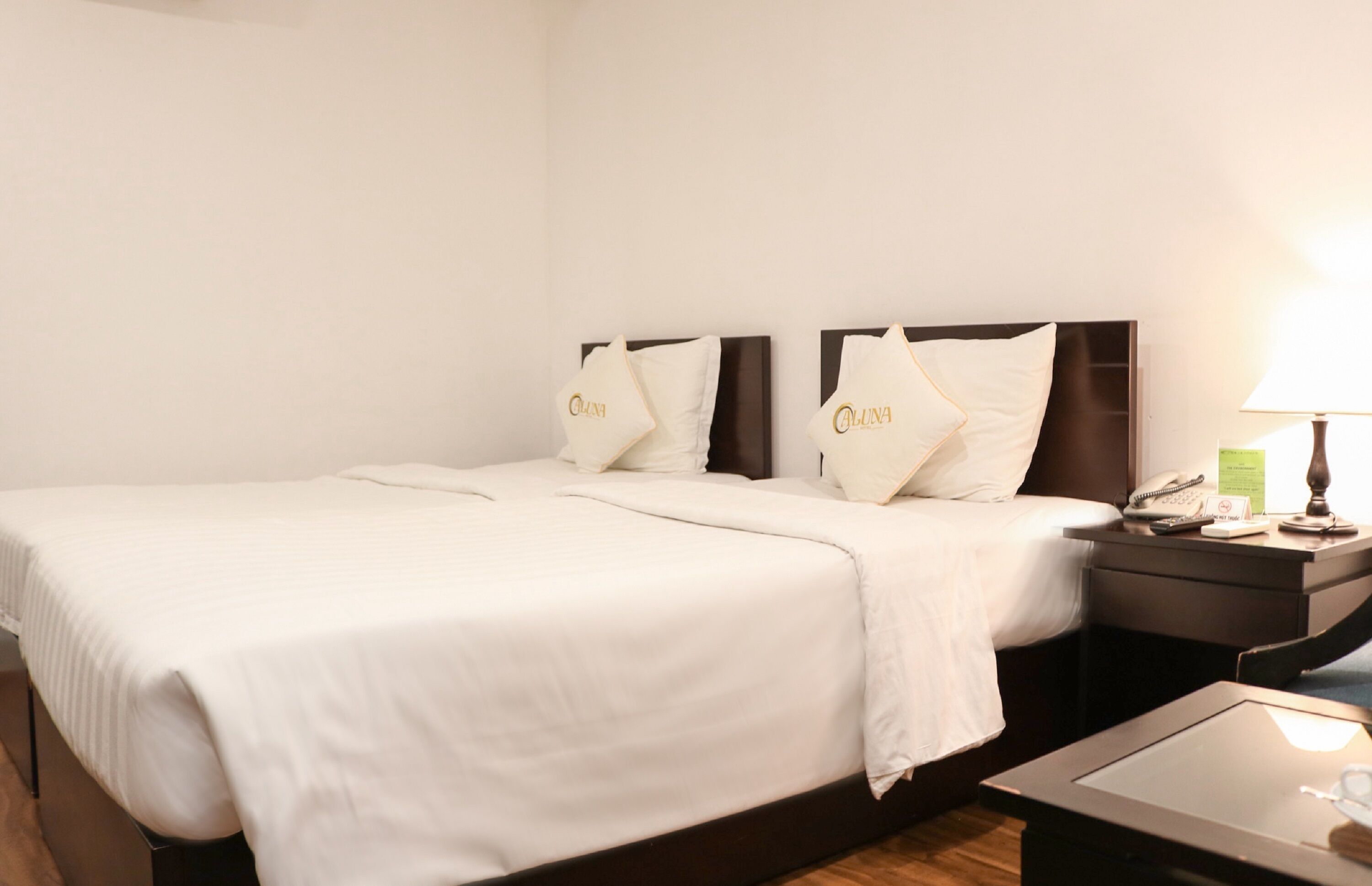 A hotel room with a large bed, two pillows with a logo, a bedside table with a lamp, and a small table with a phone and a notepad.