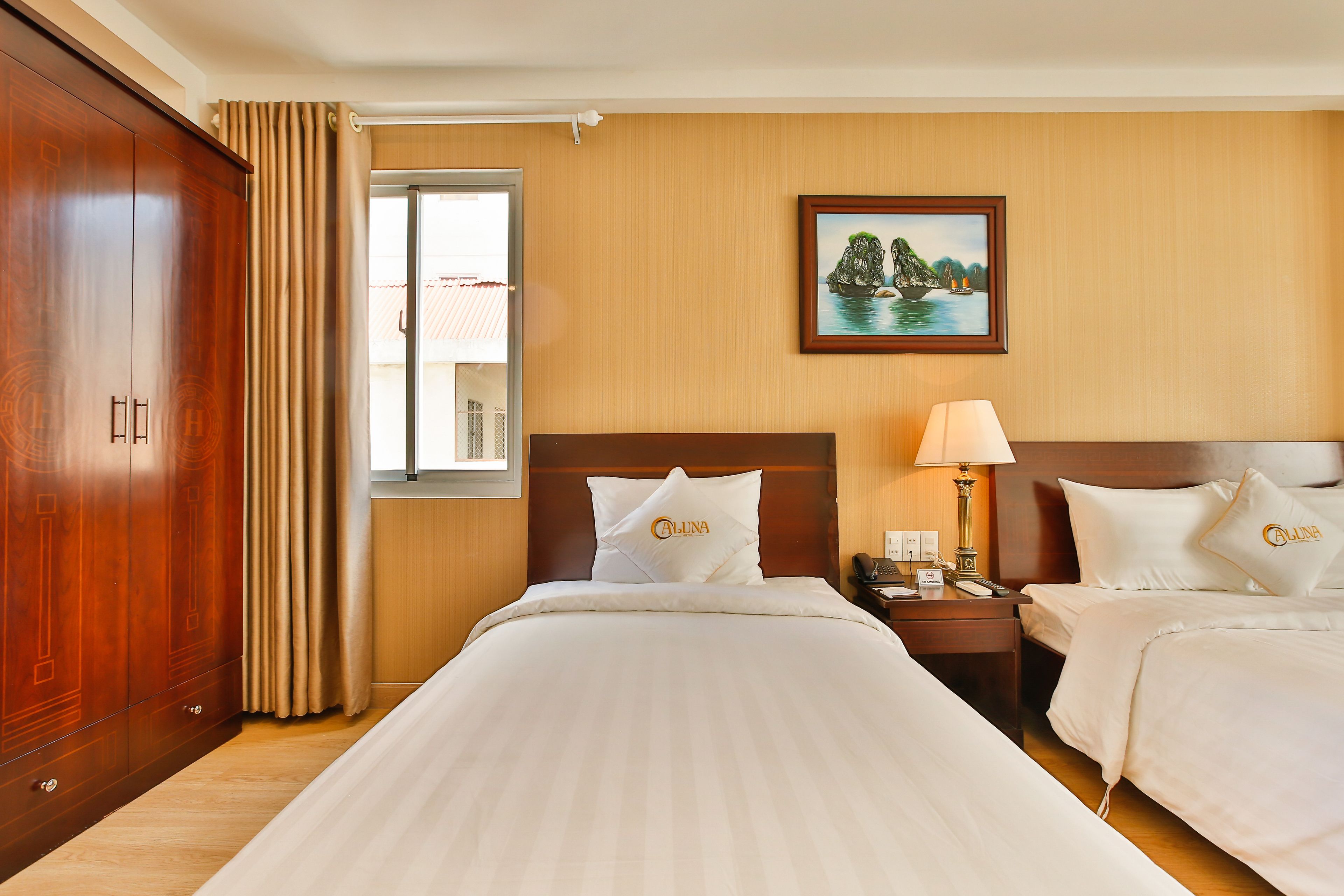 A hotel room with two beds, a large wardrobe, a window with curtains, a bedside table with a lamp, and a painting on the wall.