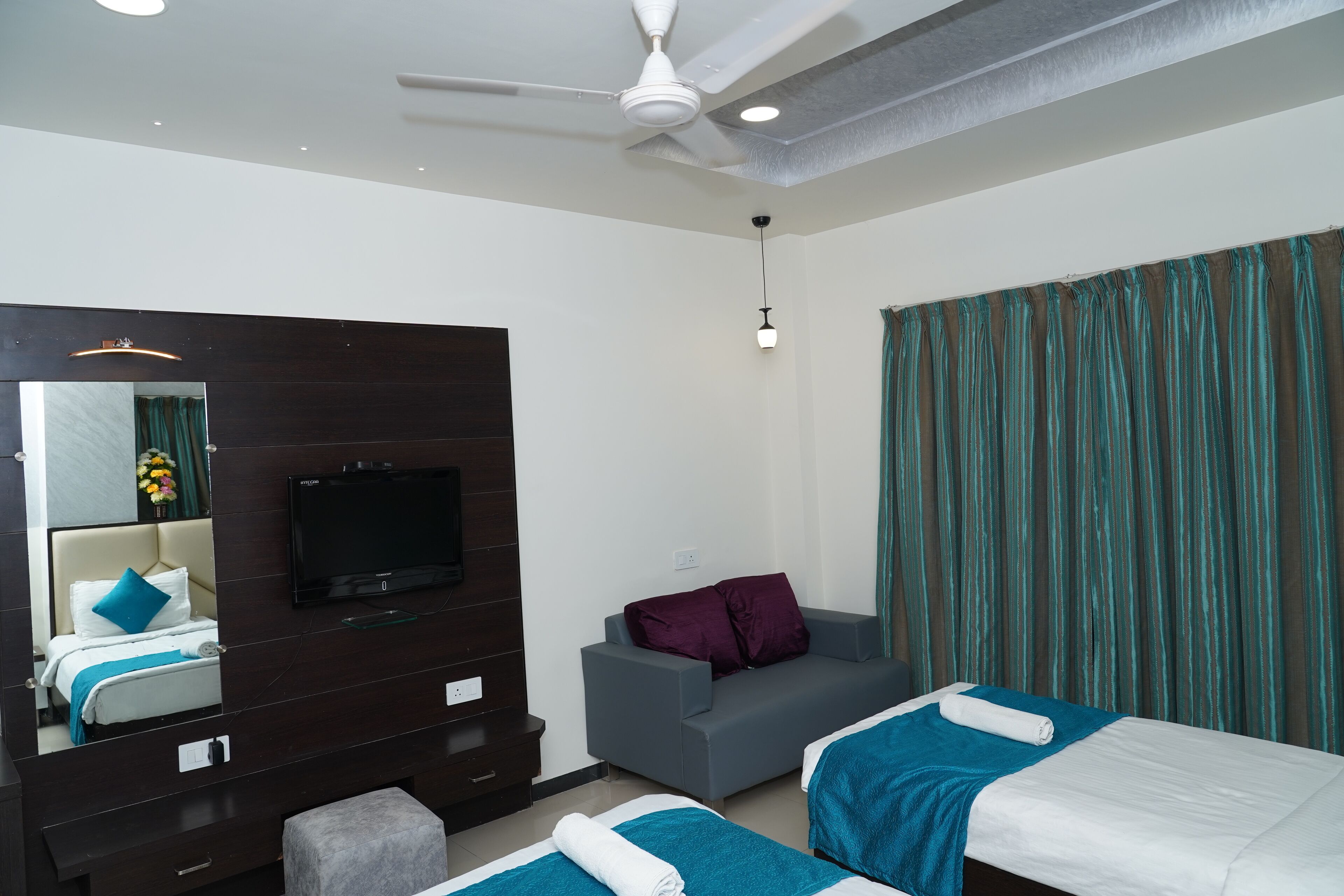 A hotel room with a bed, a sofa, a TV, a ceiling fan, and a window with curtains.