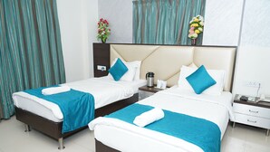 Dos camas individuales con ropa de cama blanca y azul, una mesita de noche con un teléfono y una cafetera, y un florero con flores en la pared.