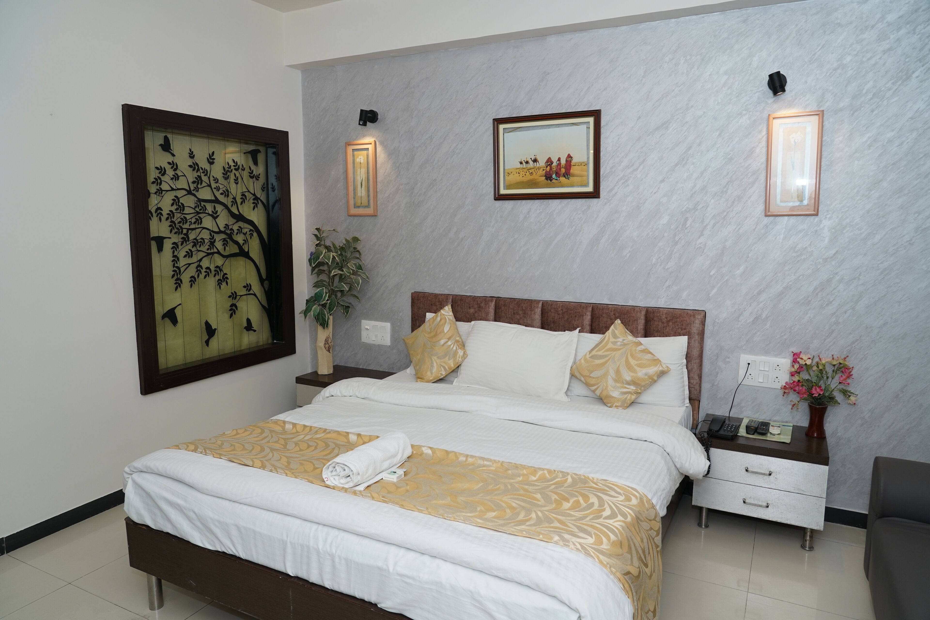 A neatly made bed with a patterned bedspread, a nightstand with a phone and a vase of flowers, and wall-mounted artwork.