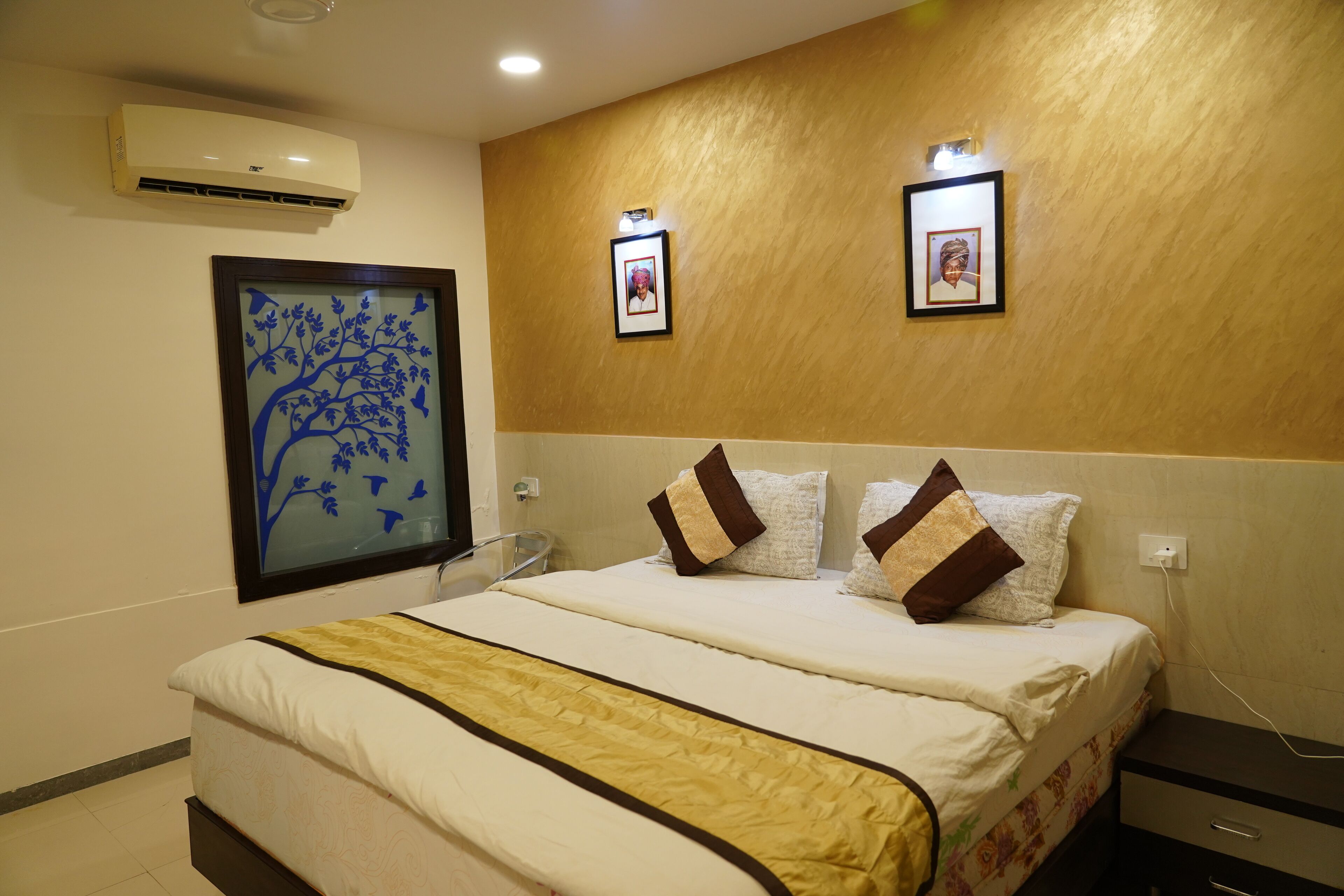 A hotel room with a bed, two wall-mounted picture frames, an air conditioning unit, and a wall-mounted light fixture.