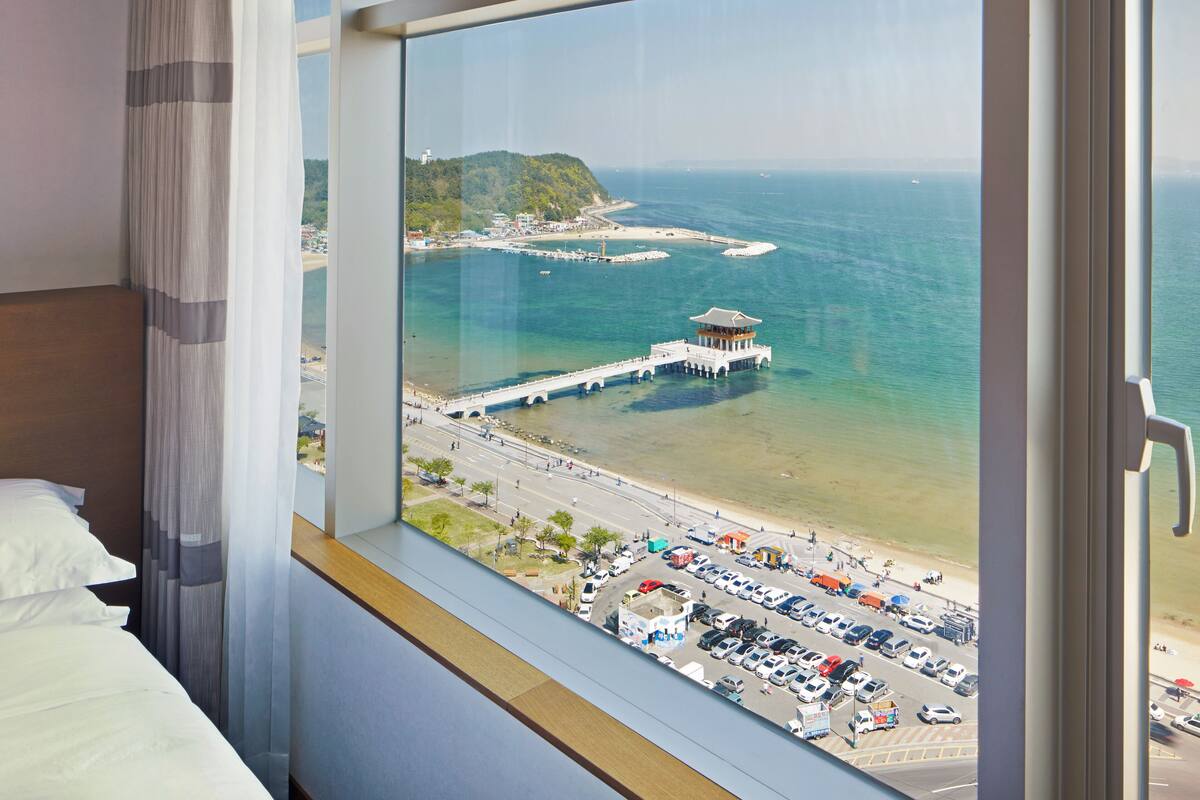 A hotel room with a large window overlooking a beach, a pier, and a mountainous landscape.