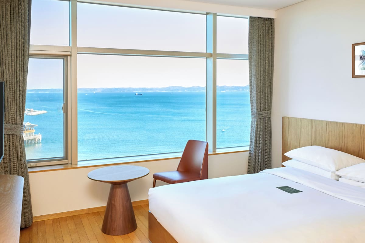 A hotel room with a large window offering a view of the sea, a bed, a chair, and a small table.