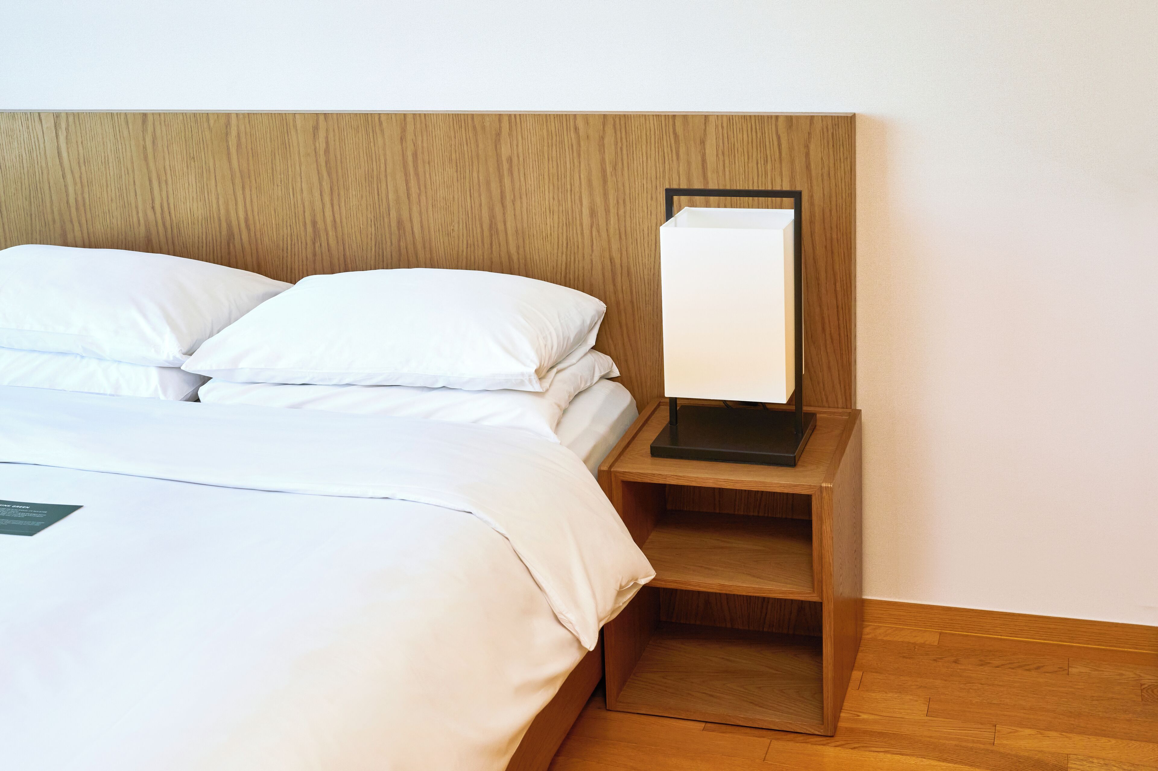 A neatly made bed with white linens and pillows, a wooden headboard, and a bedside table with a lamp.