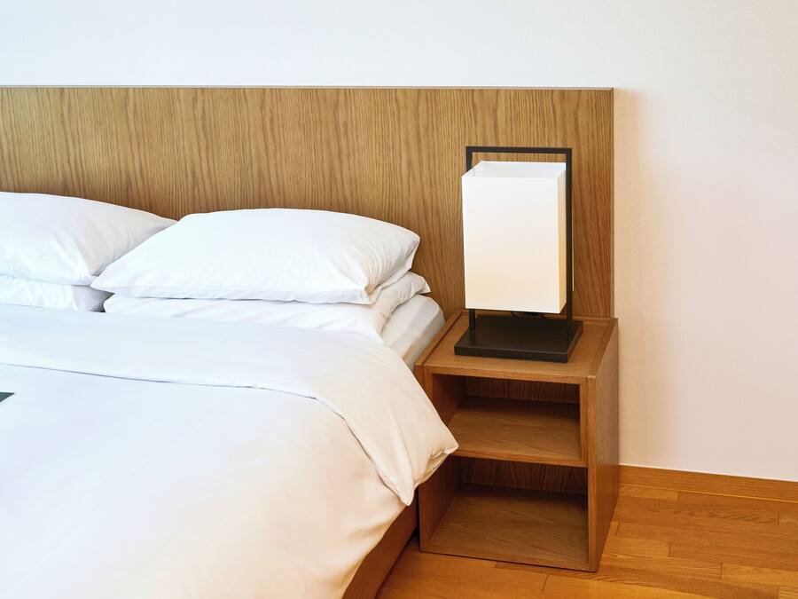 A neatly made bed with white linens and pillows, a wooden headboard, and a bedside table with a lamp.