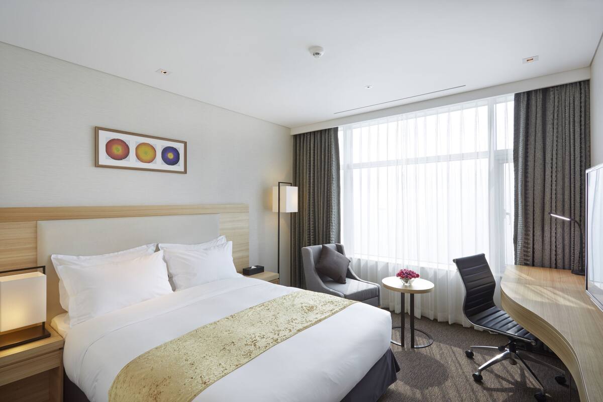 A hotel room with a large bed, a desk, a chair, a small table with a vase, and a window with curtains.