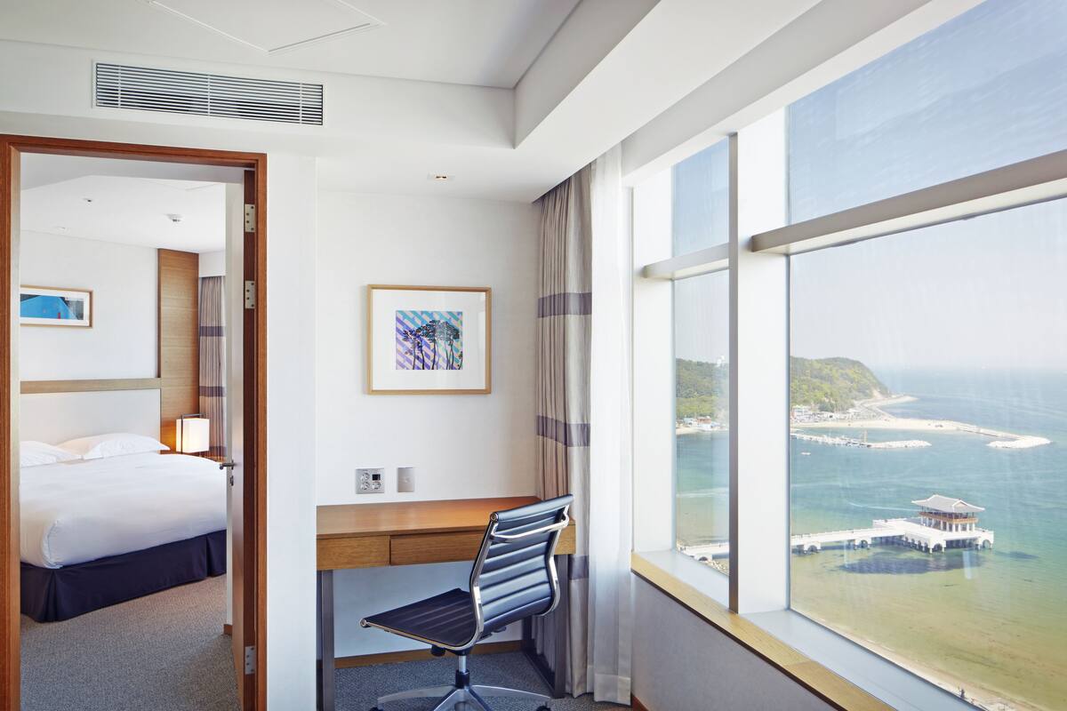 A hotel room with a bed, a desk, a chair, a large window with a view of the sea and a building, and a framed picture on the wall.