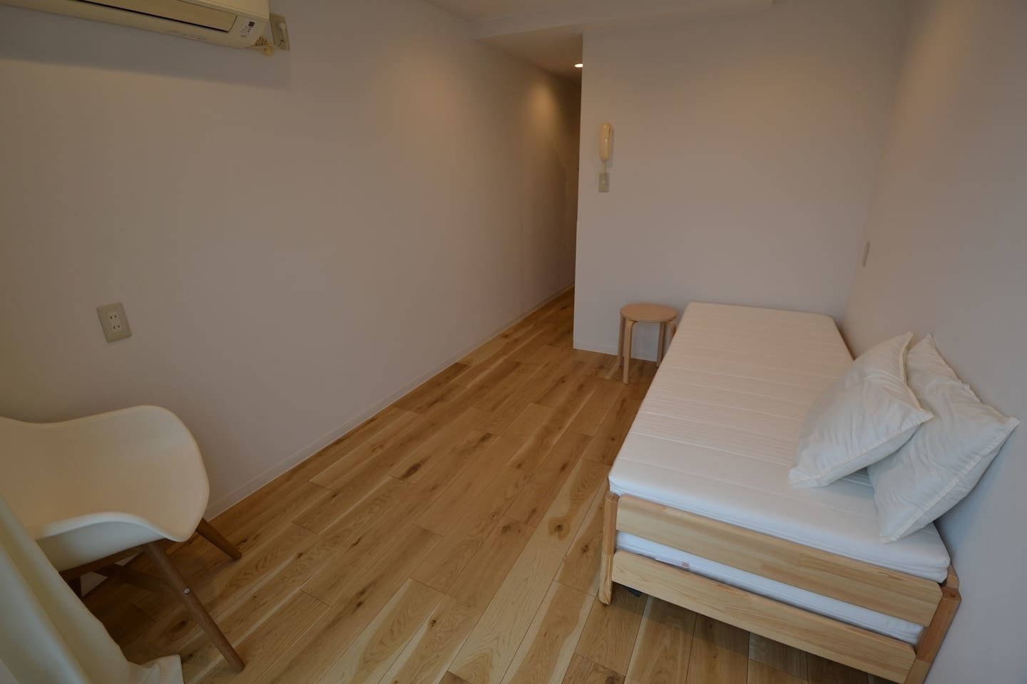 A small, sparsely furnished room with a wooden floor, a single bed with pillows, a white chair, and a small stool.