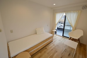A small, modern room with a single bed, a small table, and a chair. There is a window with curtains, and a balcony is visible through the window.