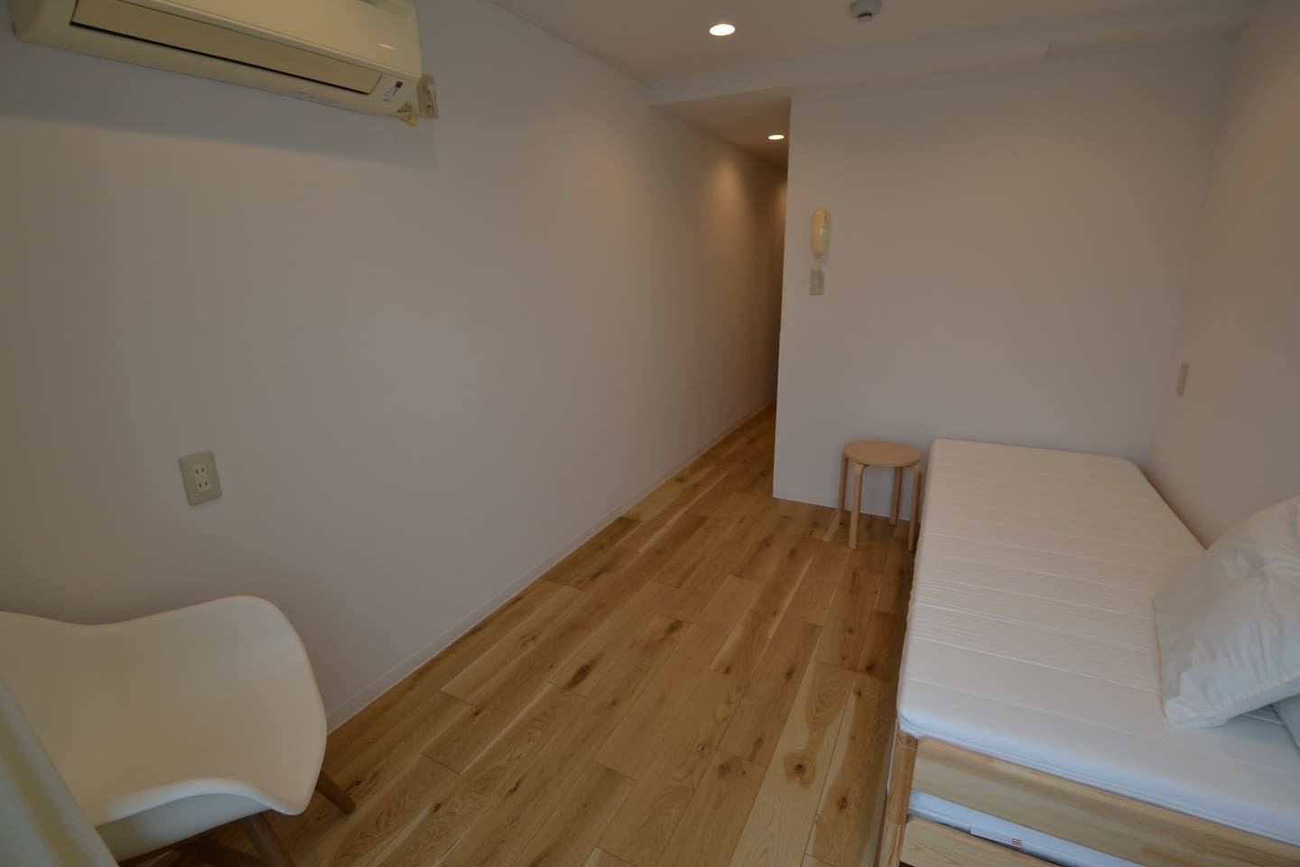 A small, sparsely furnished room with a wooden floor, a single bed, a small stool, and a white chair.