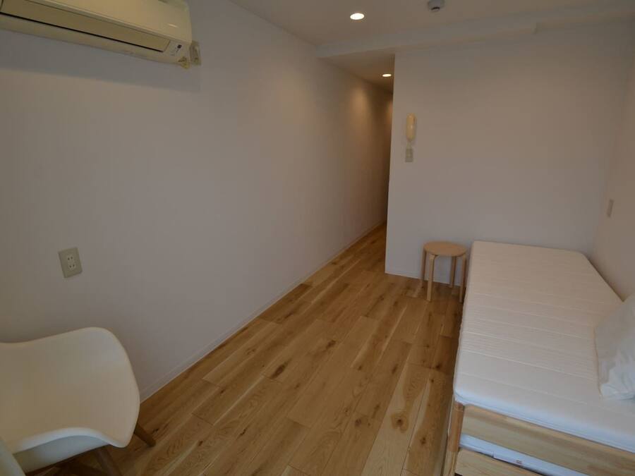 A small, sparsely furnished room with a wooden floor, a single bed, a small stool, and a white chair.