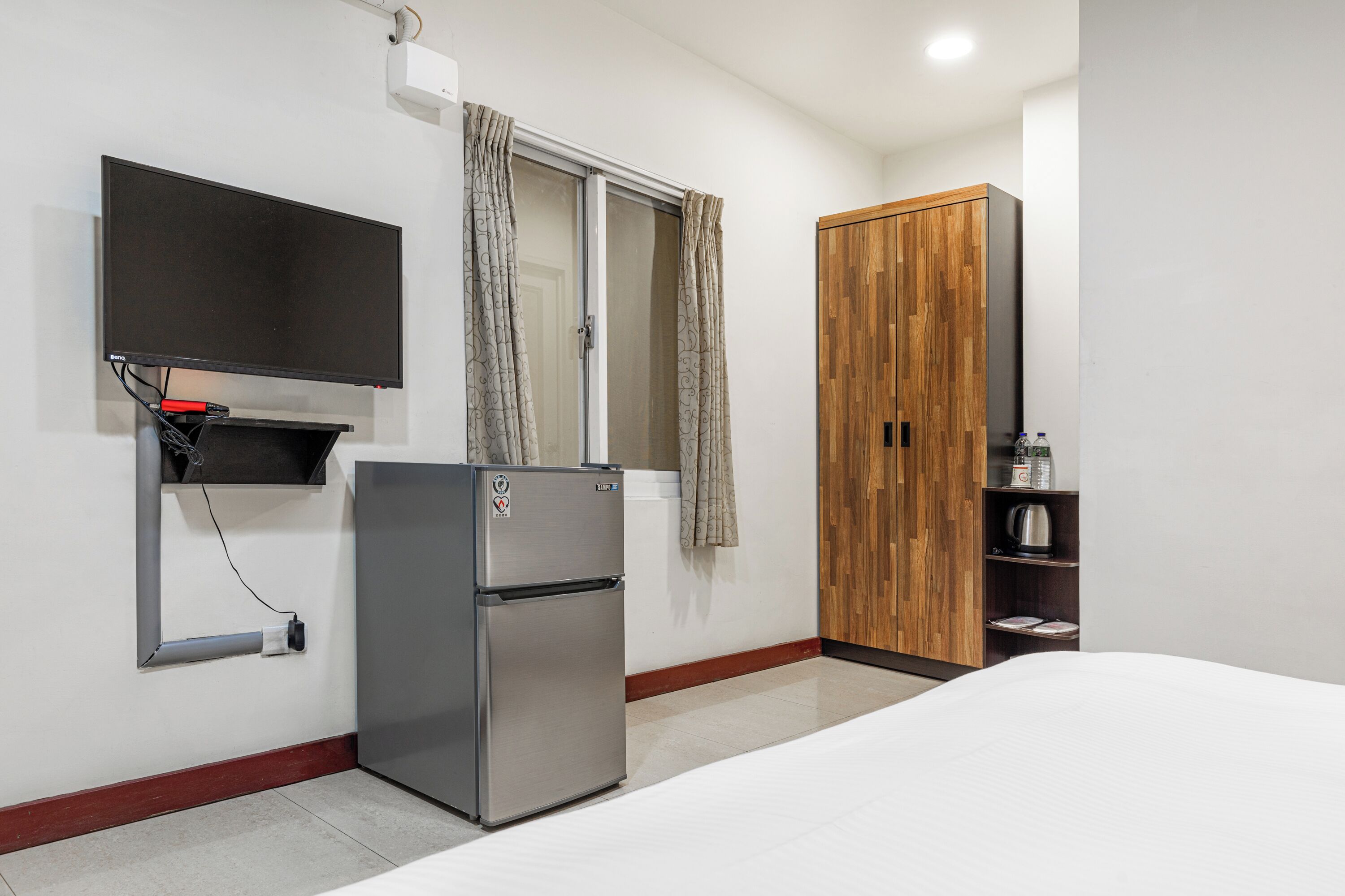 A bedroom with a bed, a refrigerator, a TV mounted on the wall, a wooden wardrobe, and a window with curtains.