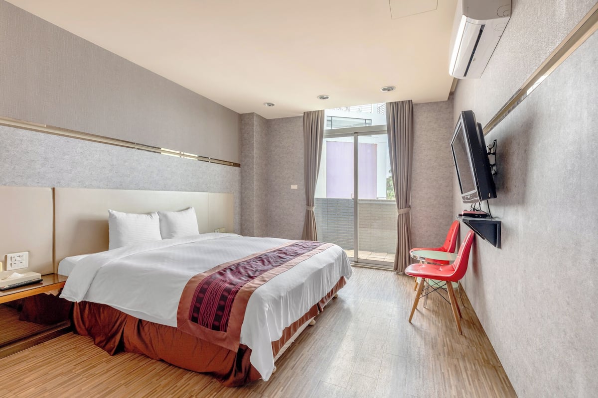 A hotel room with a large bed, a flat-screen TV mounted on the wall, a desk with a lamp, and a window with curtains.