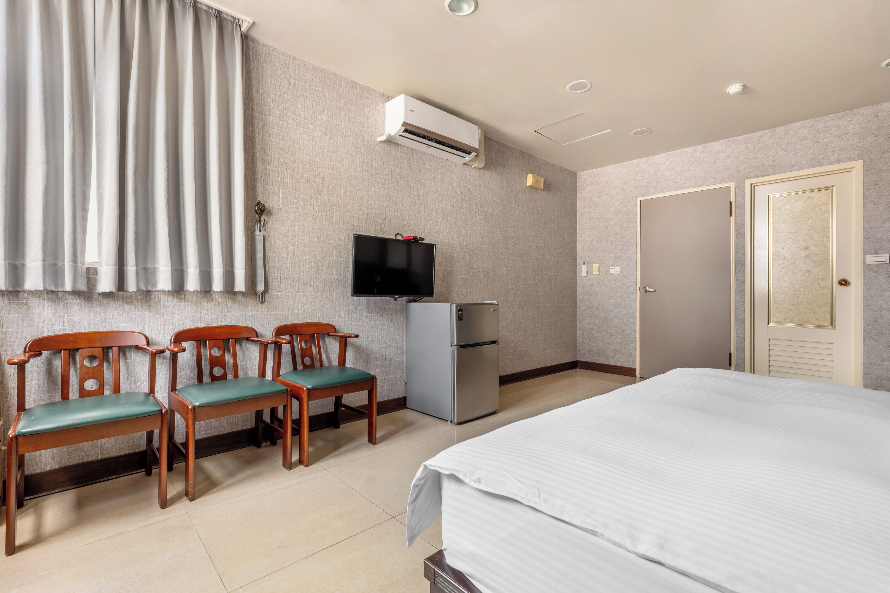 A hotel room with a bed, a TV, a refrigerator, and three wooden chairs with green cushions.