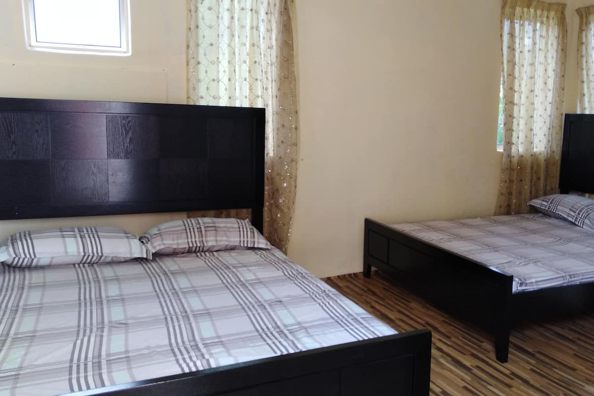 Habitación con dos camas, cada una con cabecero de madera y colcha a cuadros. El suelo es de madera y tiene una ventana con cortinas translúcidas.