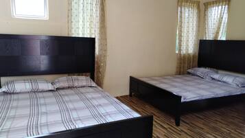 A room with two beds, each with a wooden headboard and a plaid bedspread. The room has wooden flooring and a window with sheer curtains.