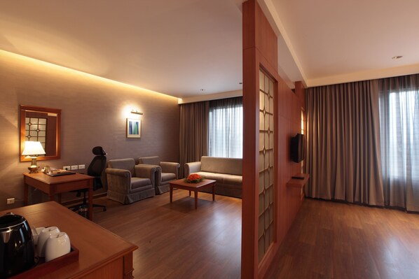 Habitación de hotel con piso de madera, un sofá, dos sillones, una mesa de centro, un escritorio con lámpara, un televisor y un ventanal con cortinas.