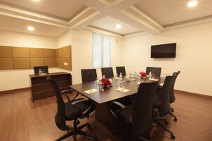 A conference room with a long table, six black chairs, a TV mounted on the wall, and a window with blinds.